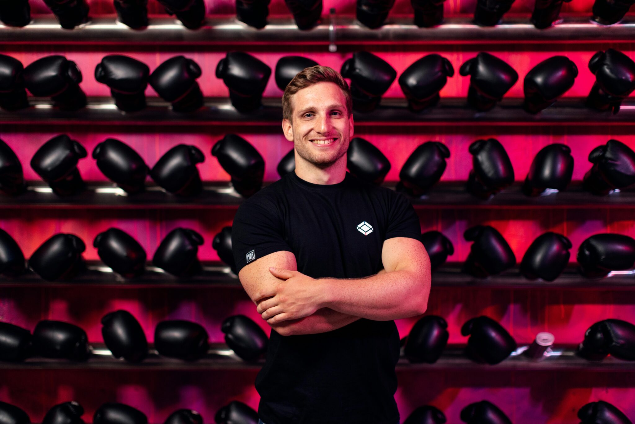 A smiling man in a gym stands confidently in front of a wall of boxing gloves.