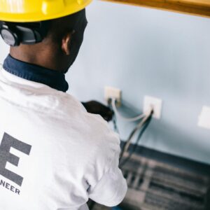 From above back view unrecognizable black field engineer wearing white shirt and protective hardhat sitting on floor and checking voltage in wall plugs